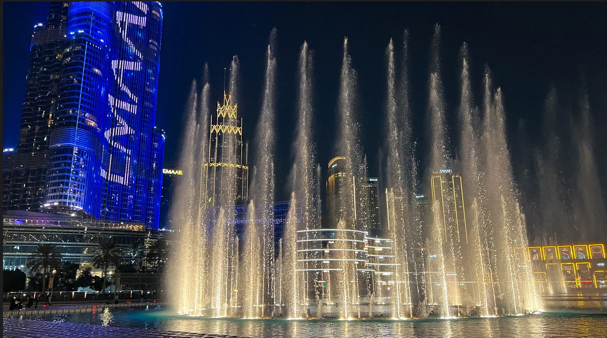 Night view of the Dubai Fountain with illuminated water jets rising from a pool against a skyline of blue lit skyscrapers and tall towers